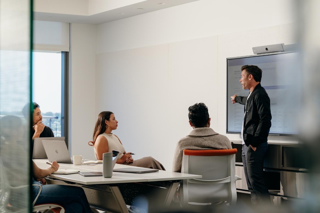 A group of diverse employees sitting in an office, a man is standing and pointing to bioinformatics results using Connected Analytics software; data is being presented on a large screen in front of a group of other employees.