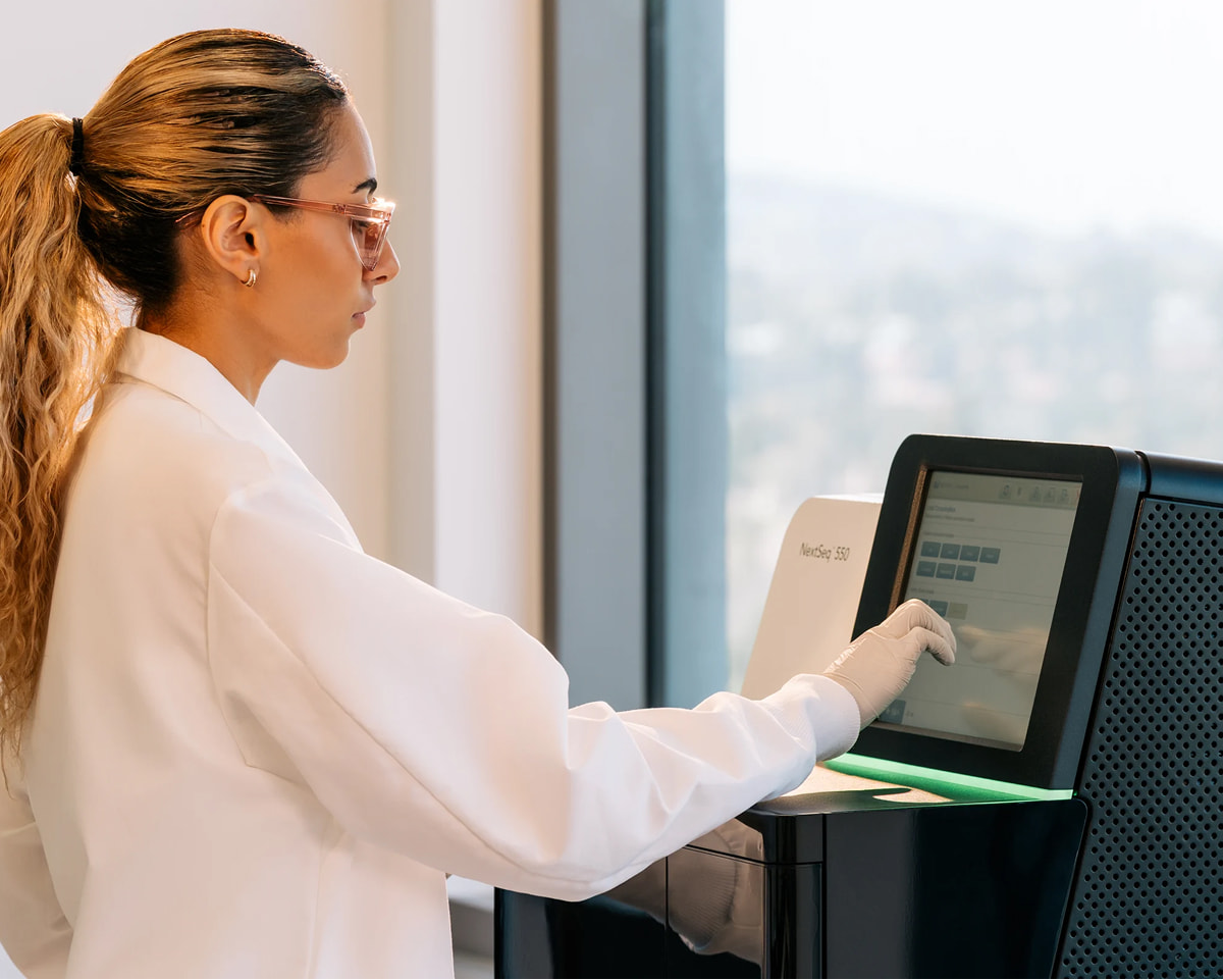 Profile view of a female scientist interacting with the load consumables touch screen on the monitor of a NextSeq 550 in front of a window; green status bar
