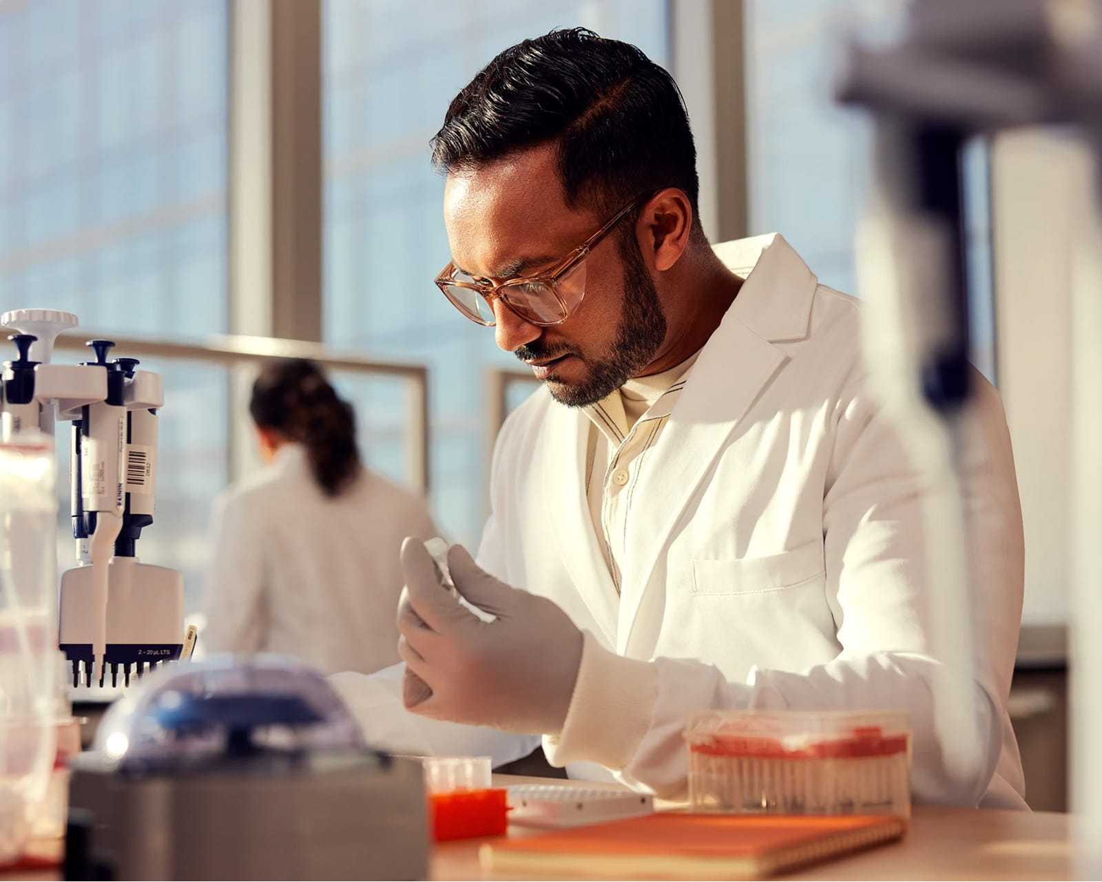 Male scientist, front side view, inspecting tube with clear liquid in wet lab, other scientist blurry in background, pipettes and plates on lab bench