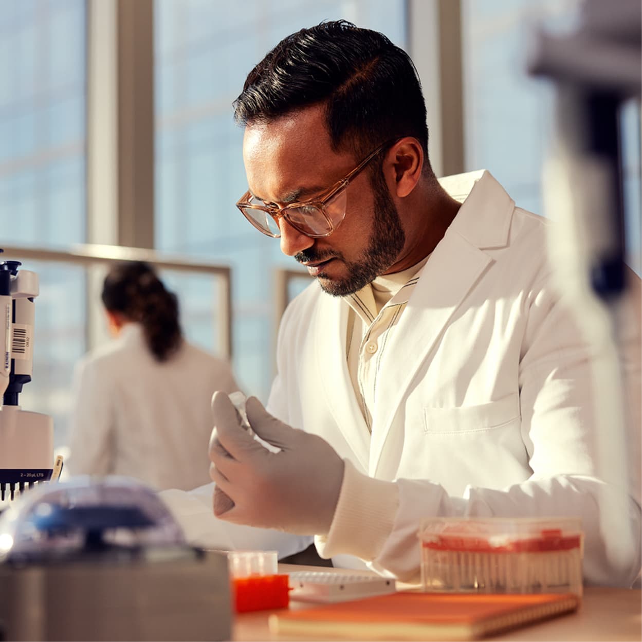 Male scientist, front side view, inspecting tube with clear liquid in wet lab, other scientist blurry in background, pipettes and plates on lab bench