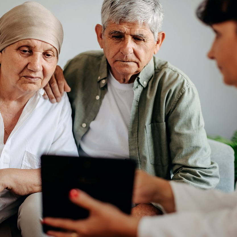 Side view of a female healthcare provider, holding a tablet, while an elderly female patient with her male partner sitting close and next to her on a couch.