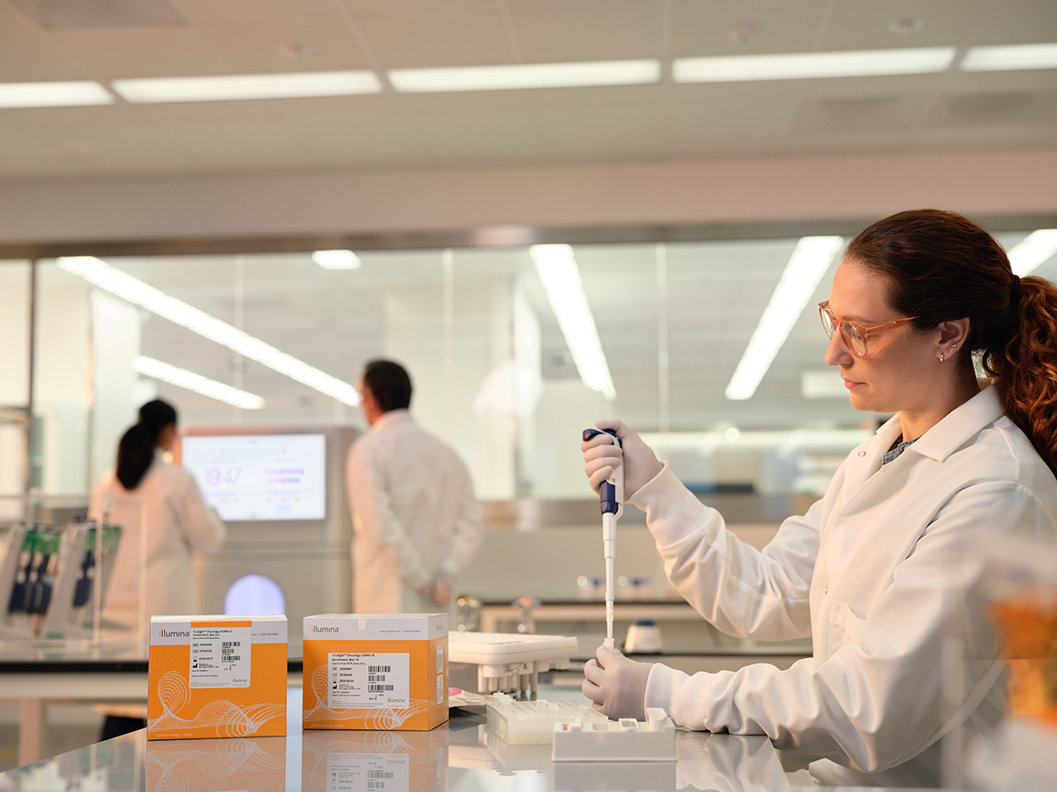 Profile image of a female scientist using a single pipette into a tube, two boxes of TruSight Oncology ctDNA v2 Enrichment library prep boxes on the lab bench with other NovaSeq X consumables; two scientists and a NovaSeq X instrument are blurry in the background.