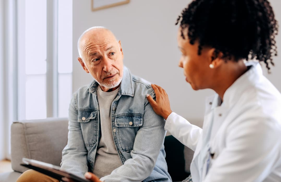 Side view of a female healthcare provider holding a tablet, looking at an elderly male patient while sitting on a couch.
