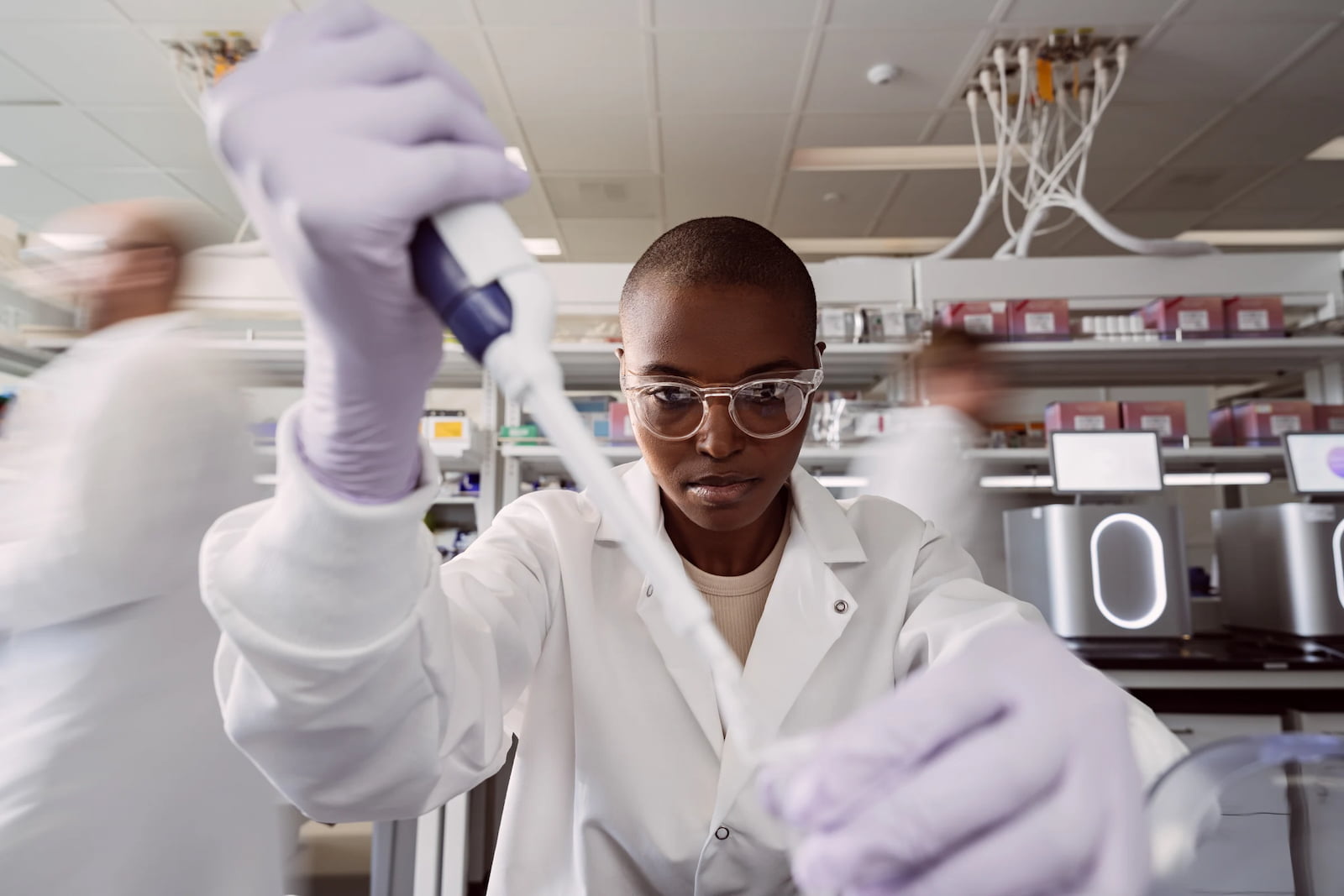 Female scientist holding a single pipette filled with clear liquid in one hand and a tube in the other; blurry scientists are moving in the background; two MiSeq i100 instruments are mid-run on the lab bench behind the scientist.