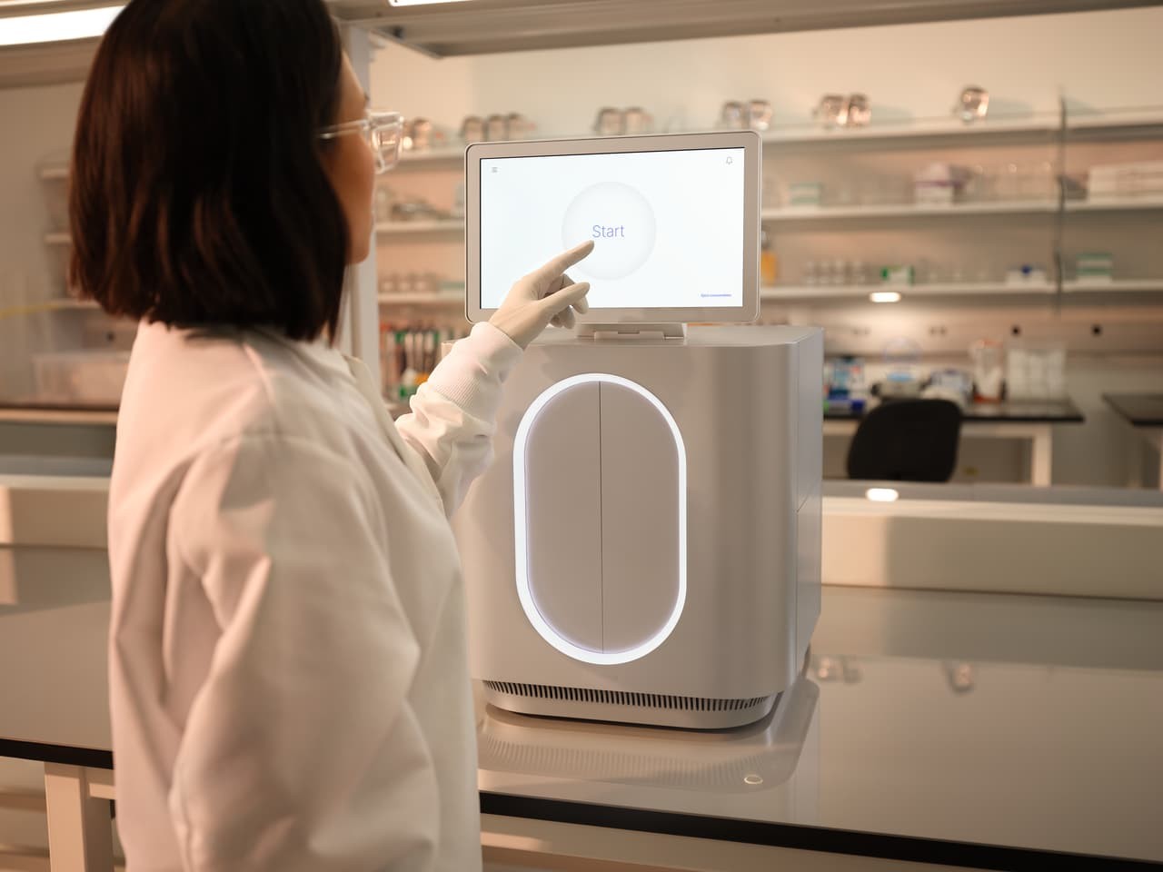 Back view of female scientist using finger to make a selection from the start touch screen monitor, front view of the MiSeq i100 on the lab bench, status bar is white.